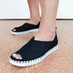 A person wearing black open-toe slip-on shoes with perforated uppers and chunky white ridged soles, standing on a speckled terracotta floor against a plain white wall.