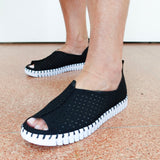 A person wearing black open-toe slip-on shoes with perforated uppers and chunky white ridged soles, standing on a speckled terracotta floor against a plain white wall.