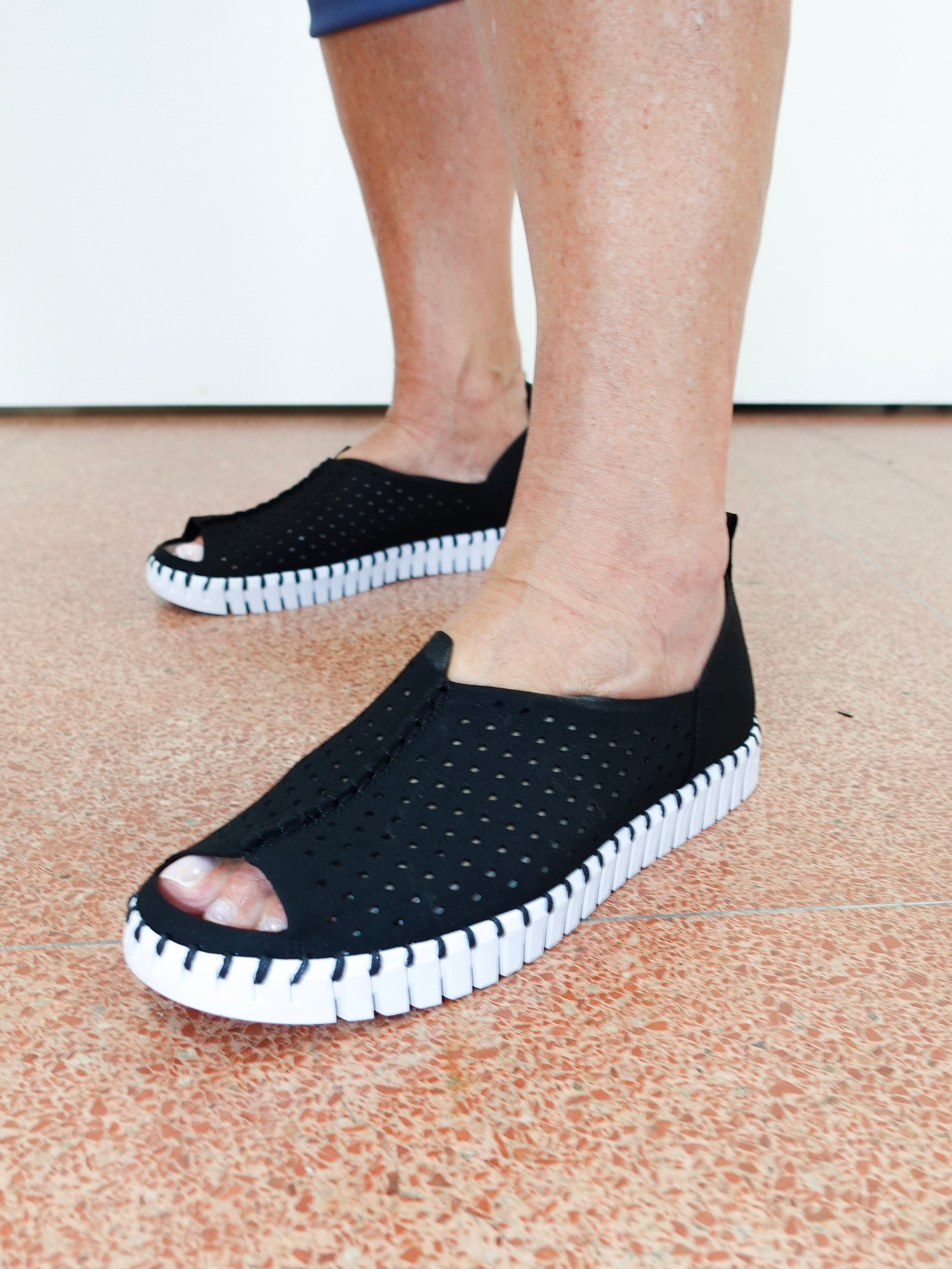 A person wearing black open-toe slip-on shoes with perforated uppers and chunky white ridged soles, standing on a speckled terracotta floor against a plain white wall.