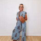 Woman wearing a blue floral jumpsuit with wide-leg pants, holding a brown leather bag against a white wall.