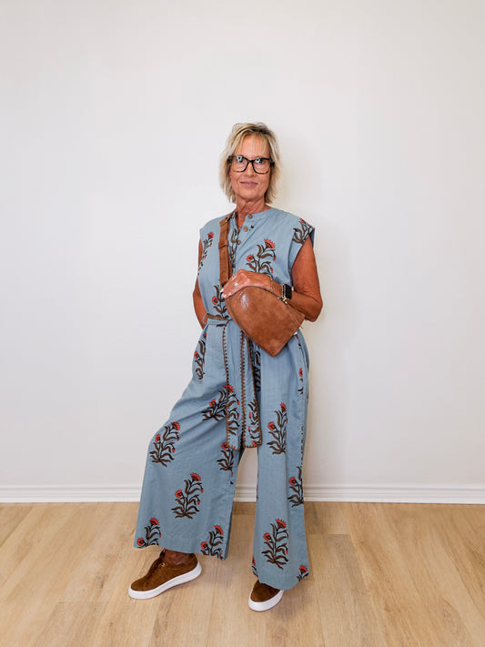 Woman wearing a blue floral jumpsuit with wide-leg pants, holding a brown leather bag against a white wall.