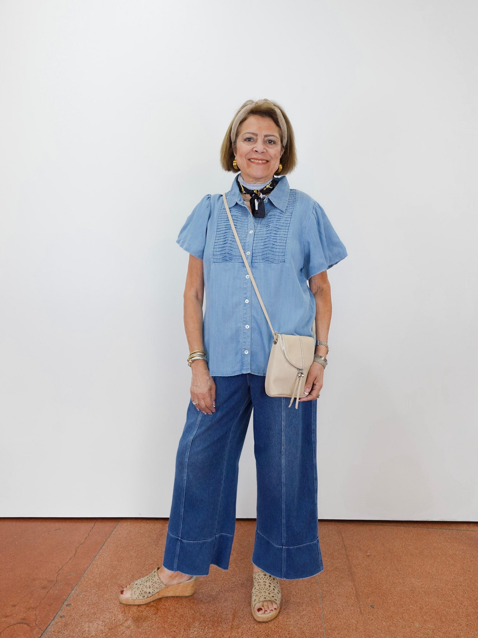 Woman wearing a blue shirt and wide-leg jeans with a beige crossbody bag against a white wall.