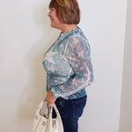Woman wearing a sheer blue floral blouse and jeans, holding a white handbag against a plain background.