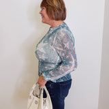 Woman wearing a sheer blue floral blouse and jeans, holding a white handbag against a plain background.