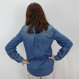 A woman viewed from behind wearing a blue denim button-up shirt and white pants with black polka dots, standing against a plain white wall.