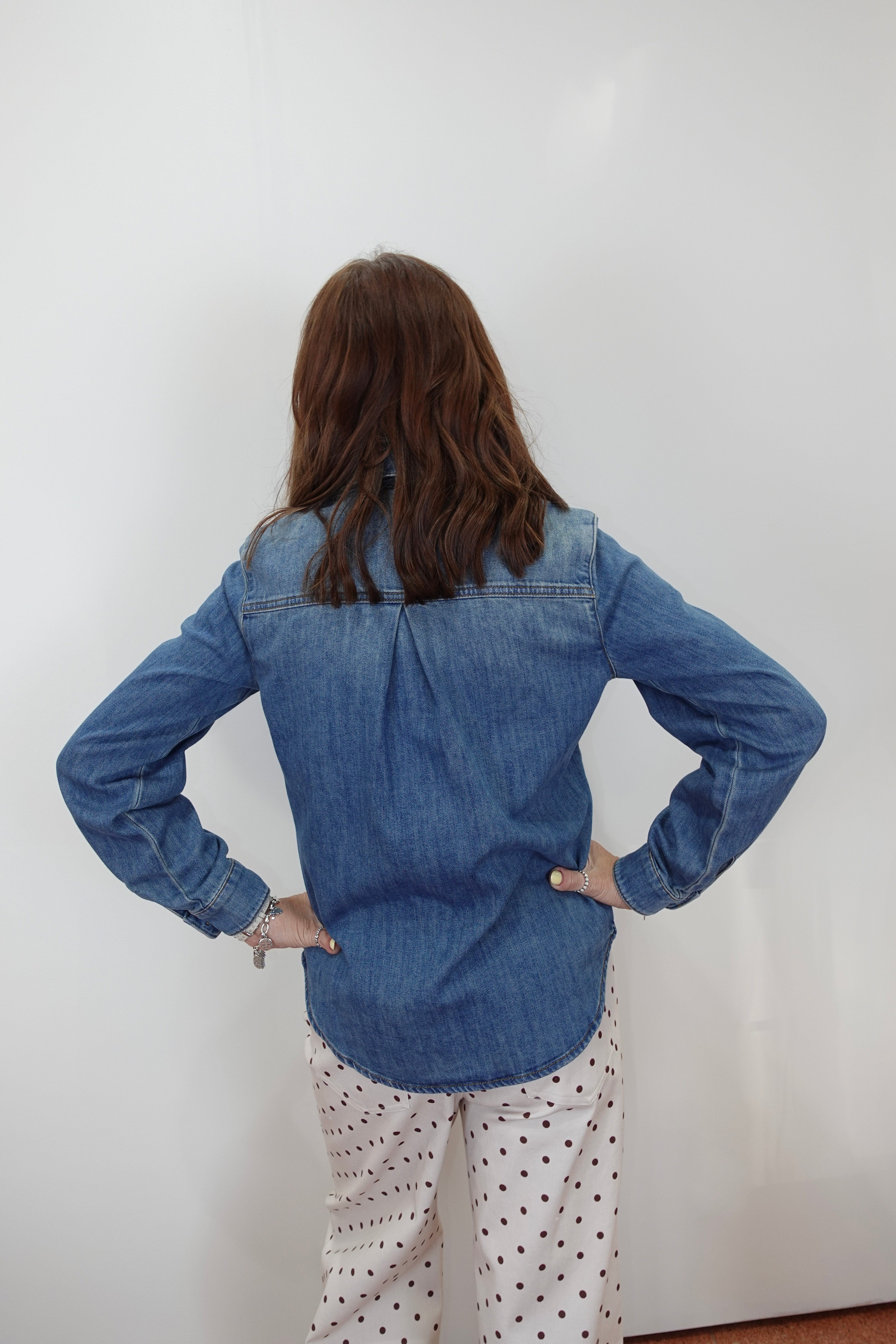 A woman viewed from behind wearing a blue denim button-up shirt and white pants with black polka dots, standing against a plain white wall.