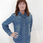 A woman with shoulder-length brown hair wearing a blue denim button-up shirt and white pants with red polka dots, standing against a plain white wall.