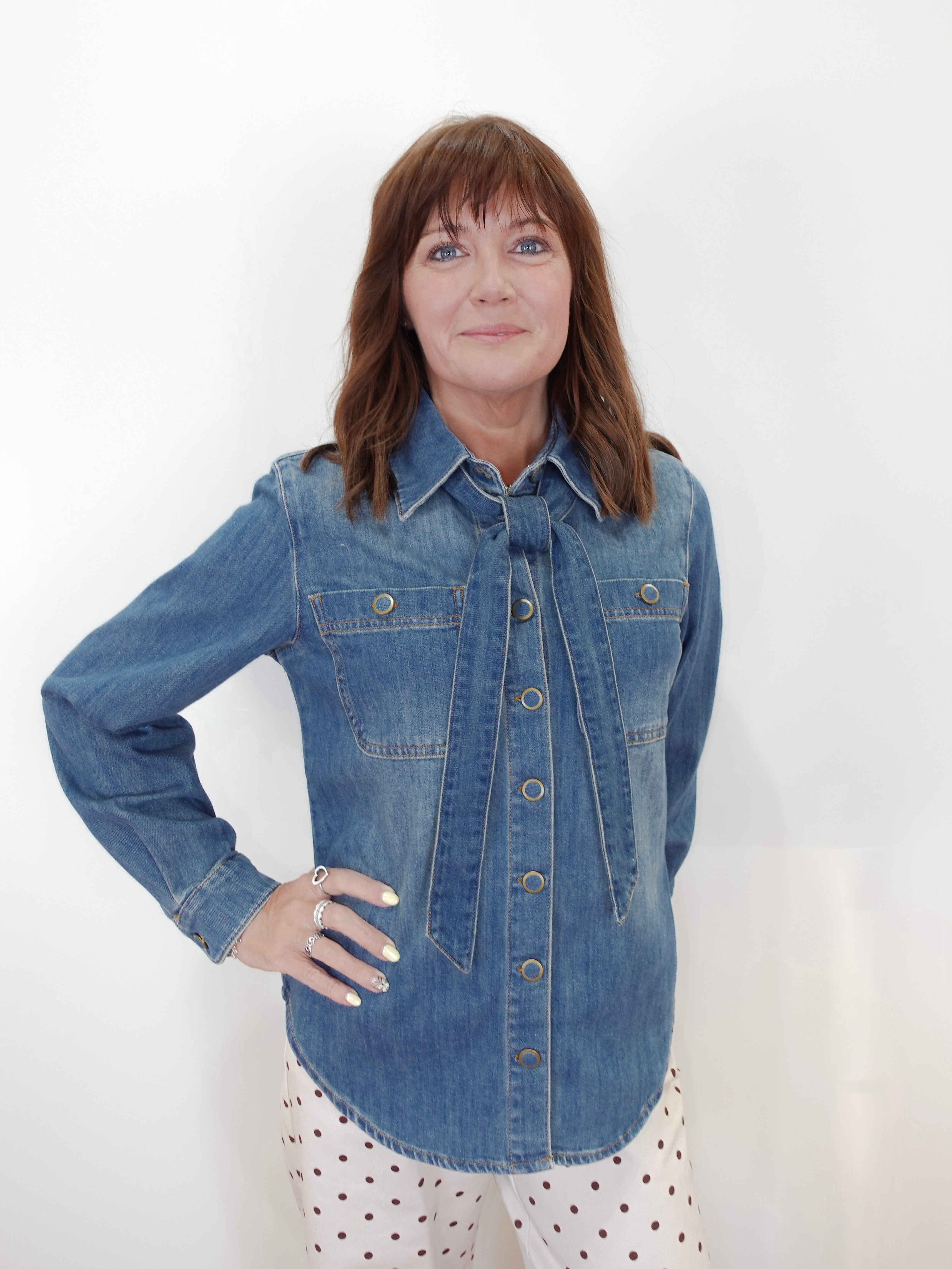 A woman with shoulder-length brown hair wearing a blue denim button-up shirt and white pants with red polka dots, standing against a plain white wall.