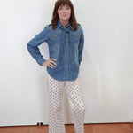 A woman stands against a plain white wall wearing a blue denim button-up shirt and white pants with black polka dots, paired with tan sandals.
