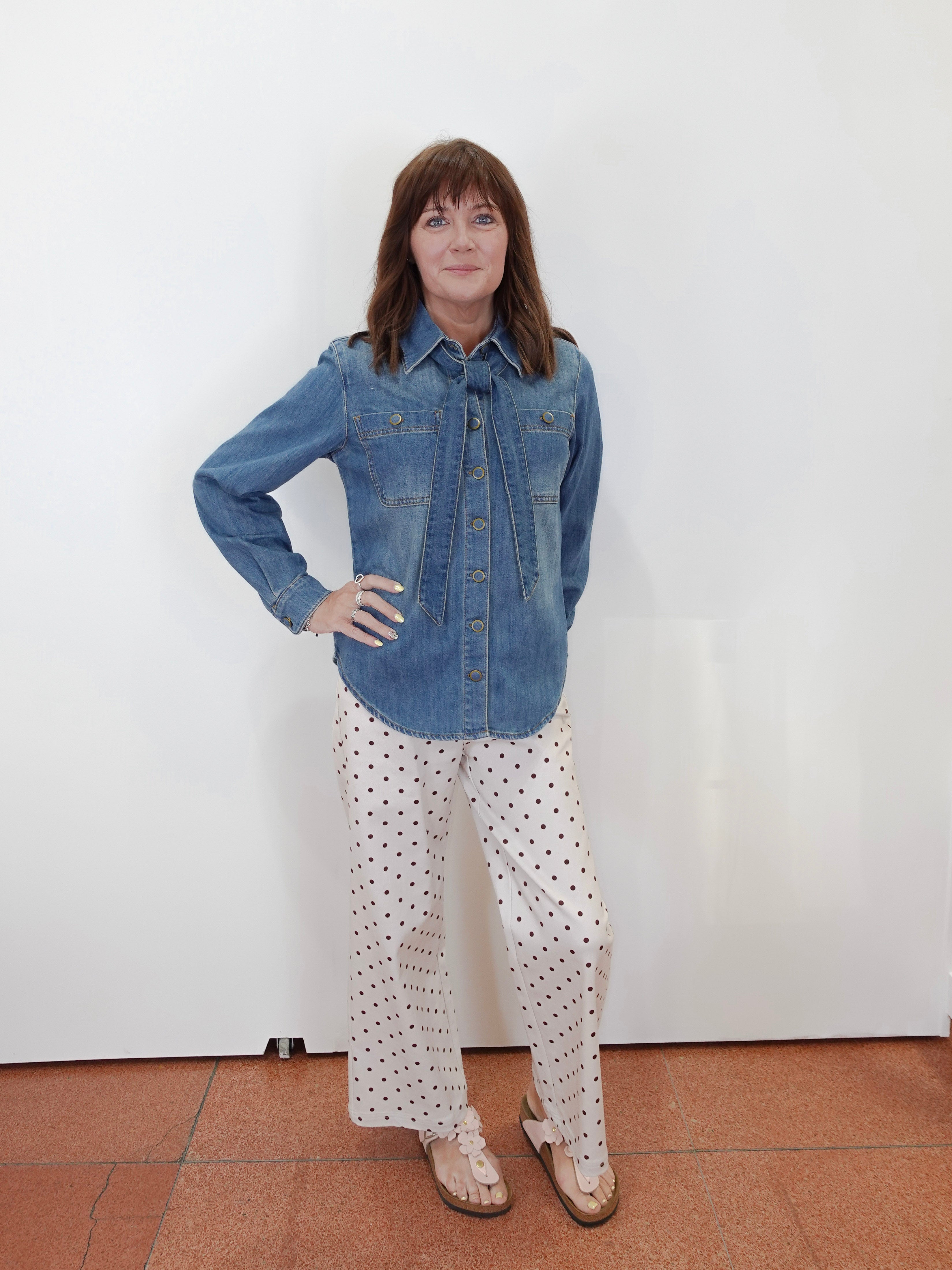 A woman stands against a plain white wall wearing a blue denim button-up shirt and white pants with black polka dots, paired with tan sandals.