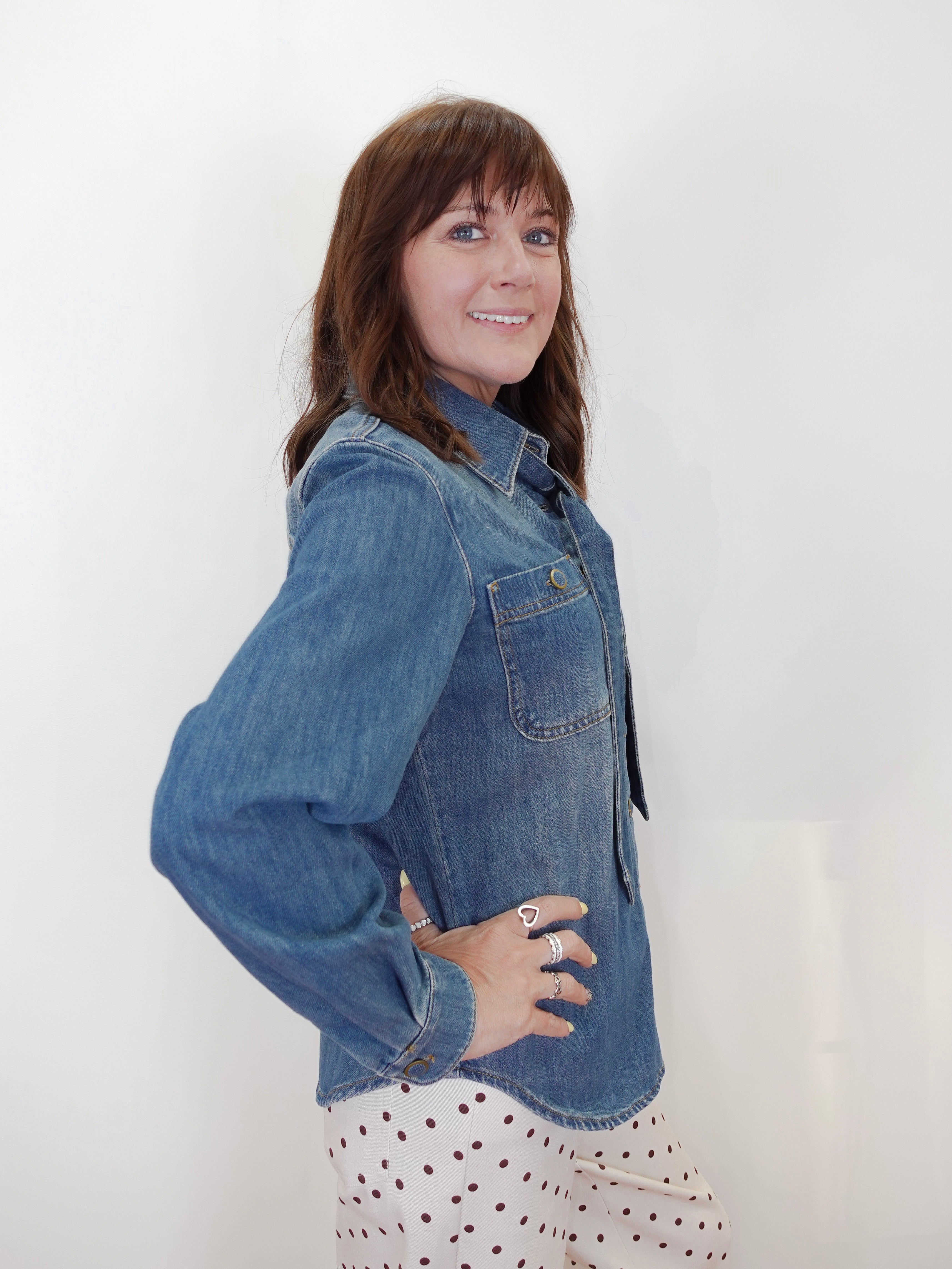 A woman with brown hair is wearing a blue denim long-sleeve button-up shirt and a white skirt with red polka dots, posing against a plain white background.