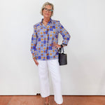 Person wearing a colorful patterned shirt and white pants, holding a black handbag against a white background.