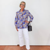 Person wearing a colorful patterned shirt and white pants, holding a black handbag against a white background.