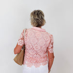 Woman wearing a pink lace blouse and white pants against a plain background