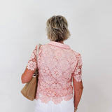 Woman wearing a pink lace blouse and white pants against a plain background