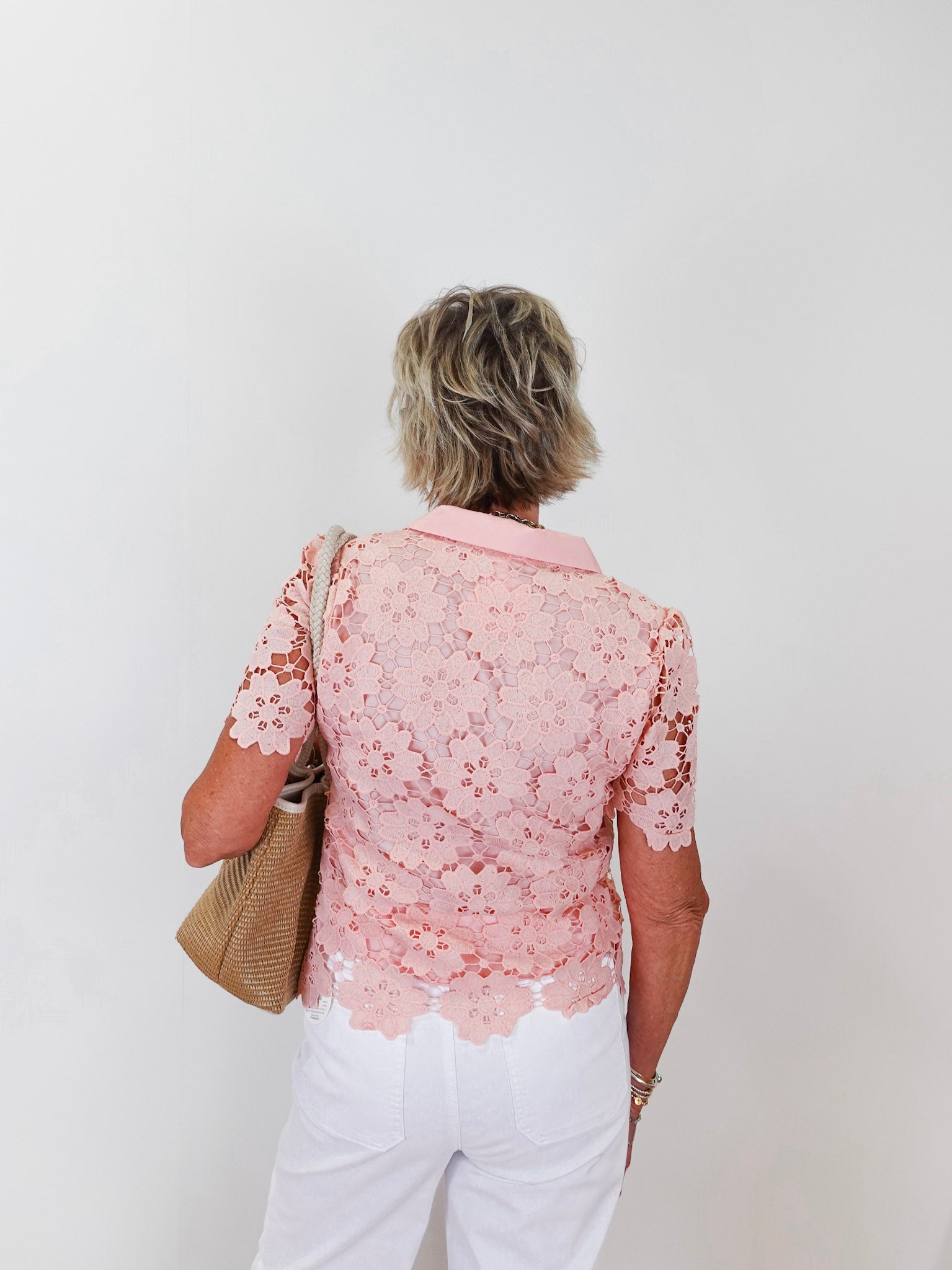 Woman wearing a pink lace blouse and white pants against a plain background