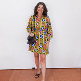 Woman wearing a colorful patterned dress standing against a white wall.