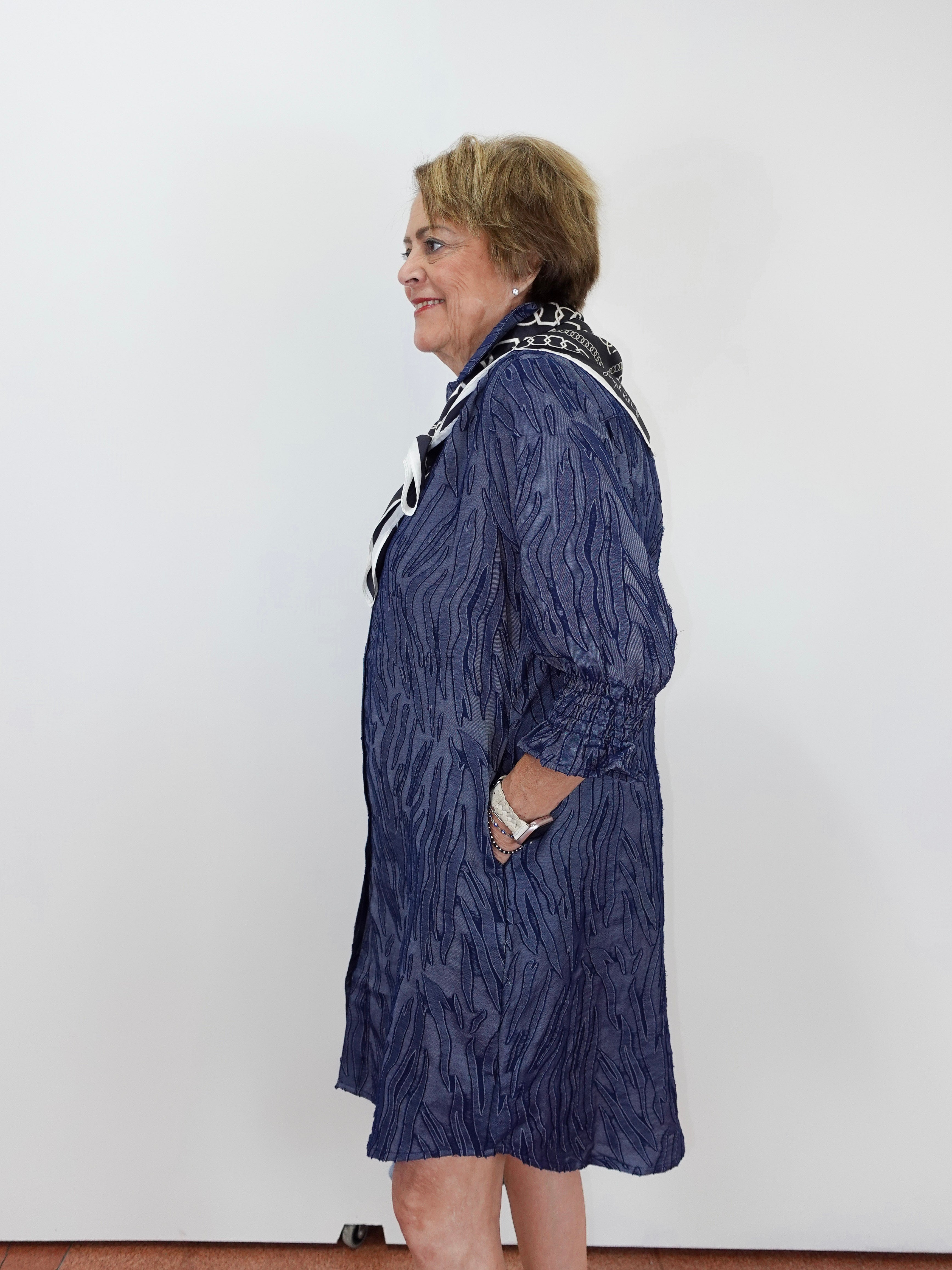 A woman wearing a textured navy blue longline cardigan with black and white patterned trim, standing in profile against a plain white wall.