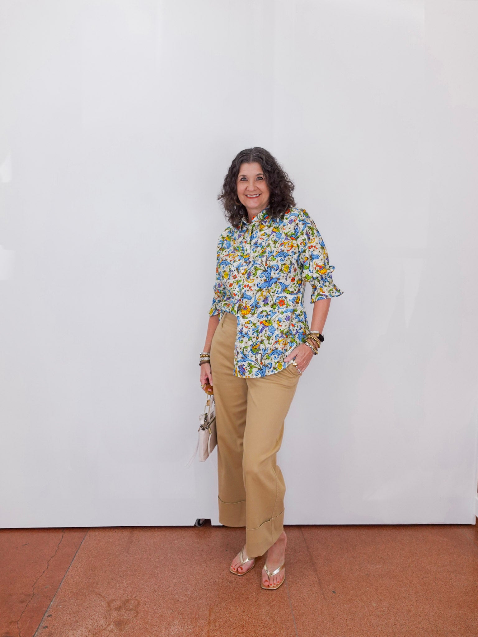 Woman wearing a floral blouse and beige pants against a white wall.