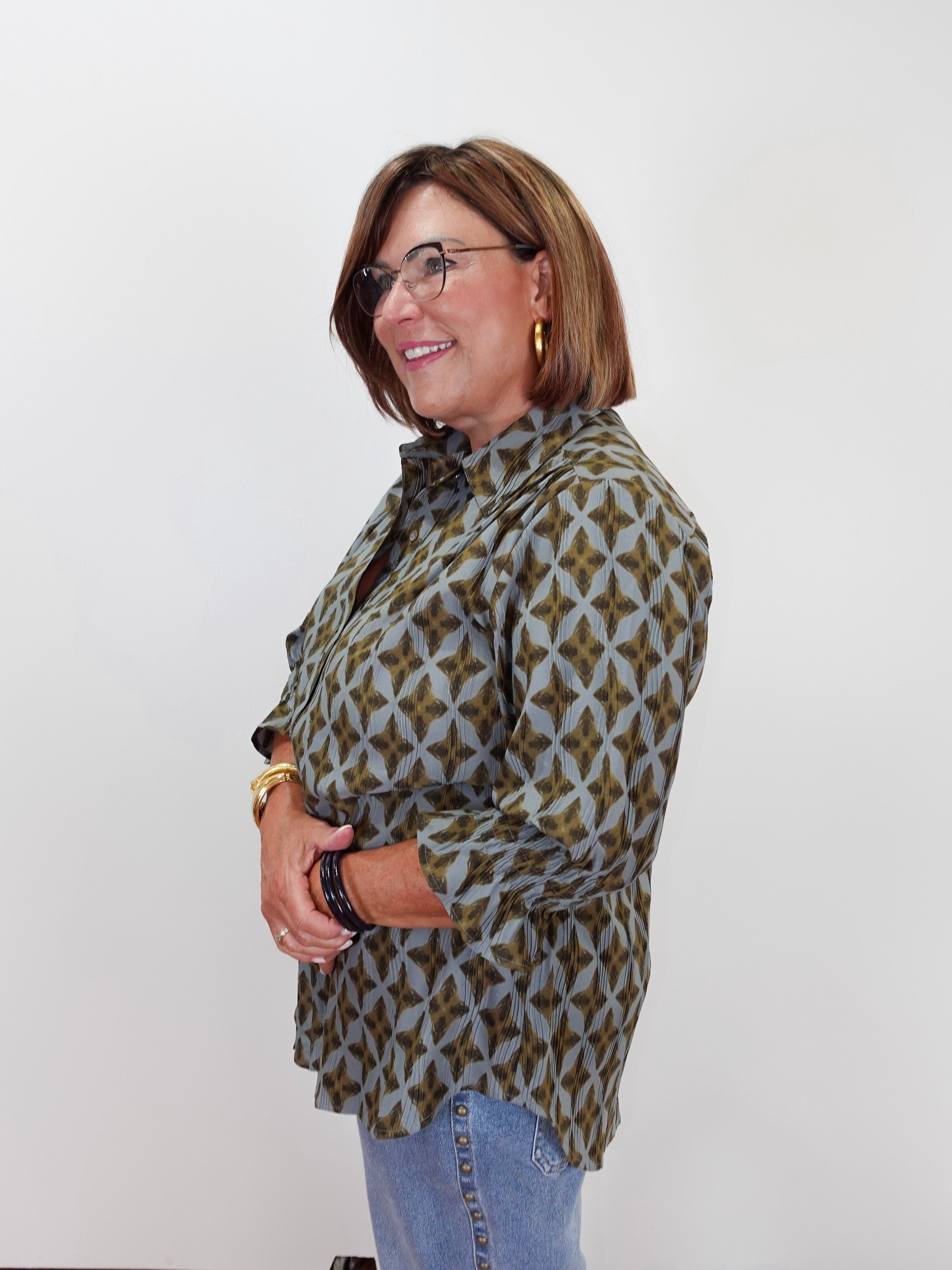 A woman with glasses and brown hair is shown from the waist up, wearing a long-sleeve button-up shirt with a brown and gray geometric pattern, paired with light blue jeans, against a plain white background.