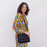 Woman wearing a colorful patterned dress holding a black handbag against a white background
