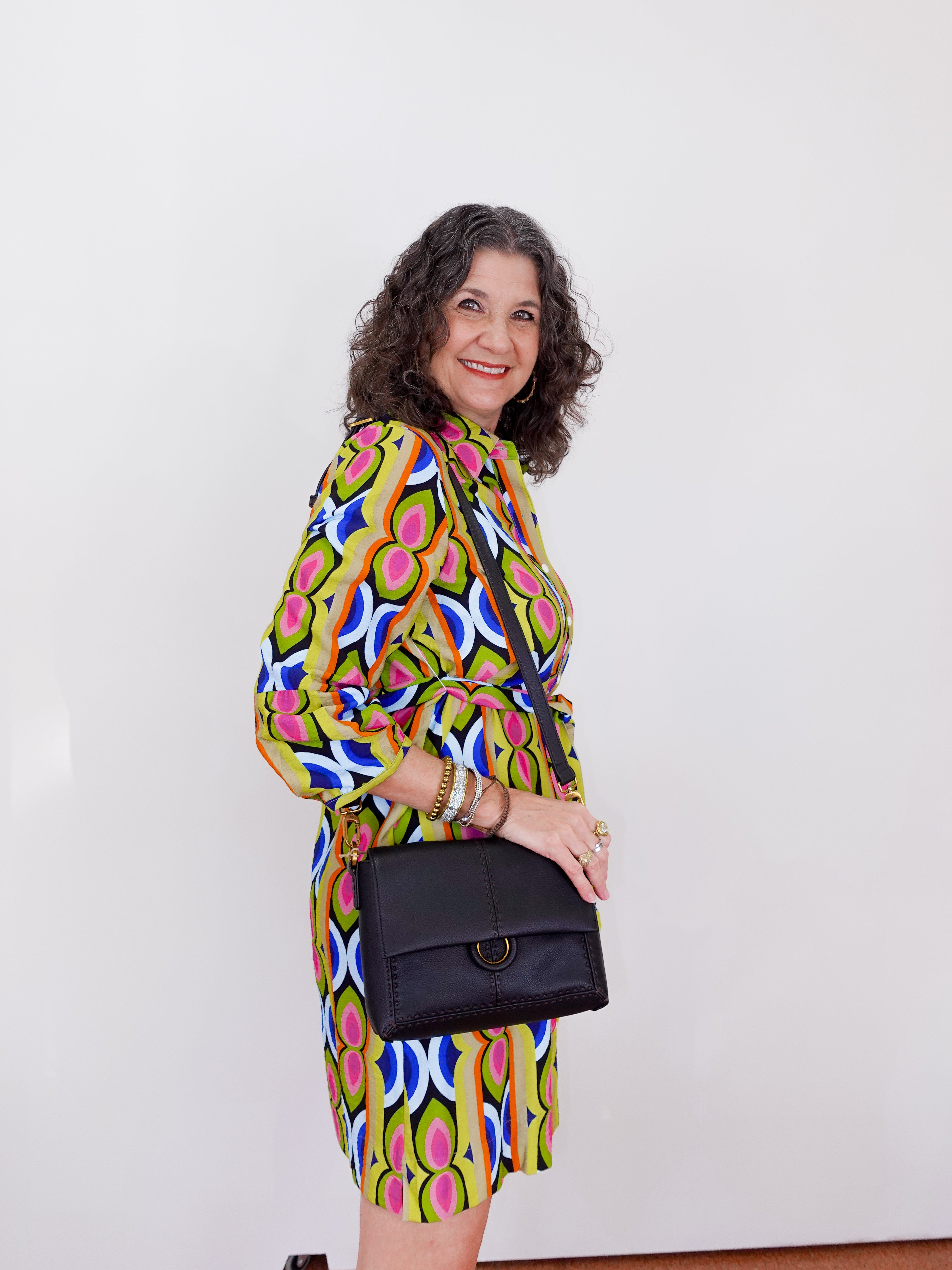 Woman wearing a colorful patterned dress holding a black handbag against a white background