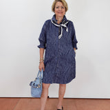 A woman stands against a plain white wall wearing a navy blue patterned shirt dress with a white and navy neck scarf, holding a light blue handbag, and wearing black platform sandals.