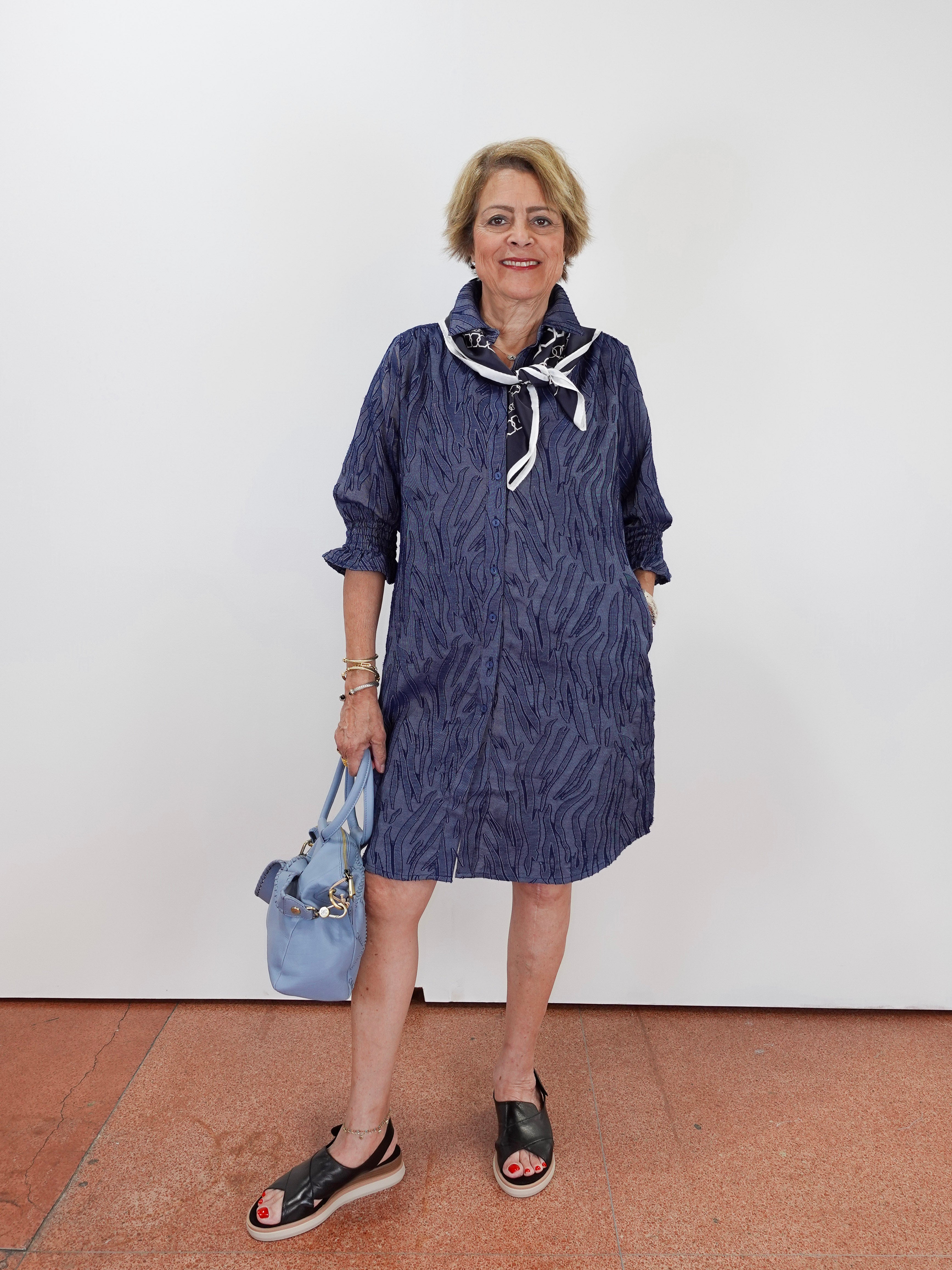 A woman stands against a plain white wall wearing a navy blue patterned shirt dress with a white and navy neck scarf, holding a light blue handbag, and wearing black platform sandals.