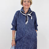 An older woman modeling a textured navy blue shirt dress with a white and navy neck scarf, holding a light blue handbag against a plain white background.