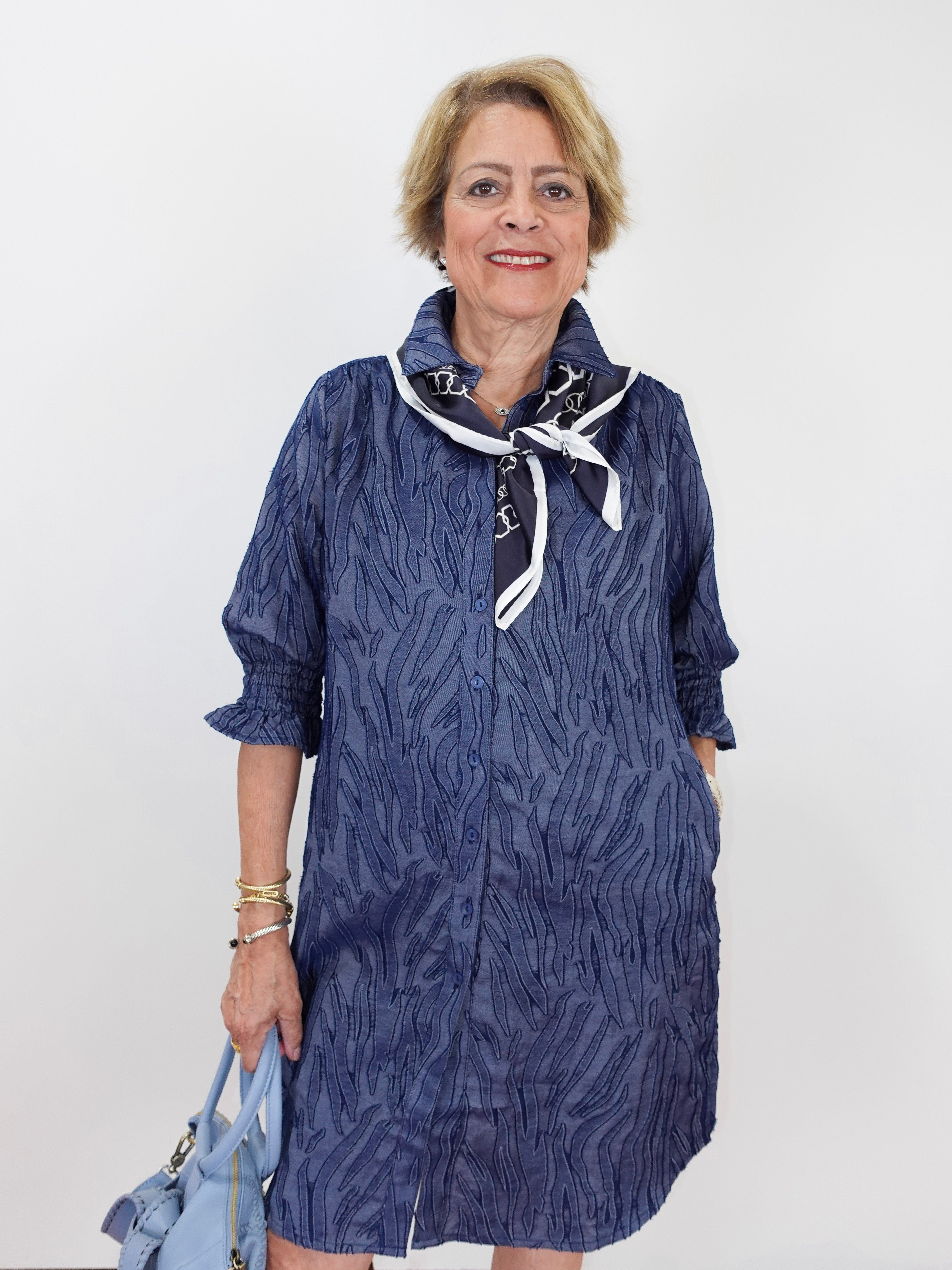 An older woman modeling a textured navy blue shirt dress with a white and navy neck scarf, holding a light blue handbag against a plain white background.