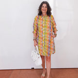 Woman wearing a colorful patterned dress holding a white handbag indoors.