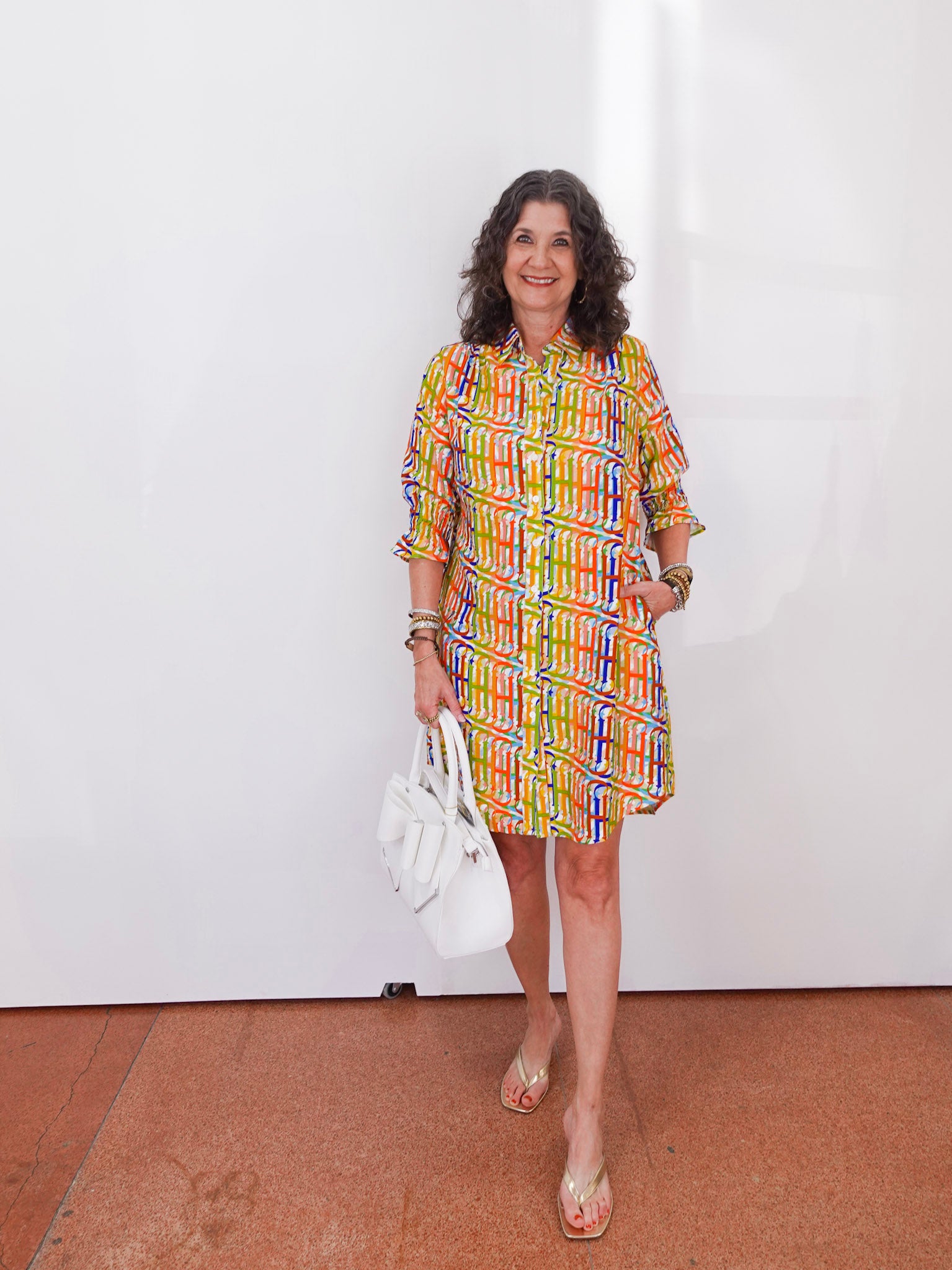 Woman wearing a colorful patterned dress holding a white handbag indoors.