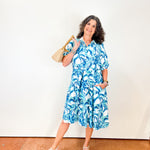 Woman wearing a blue floral dress holding a beige handbag against a white wall.