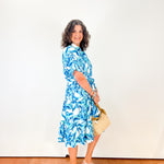 Woman in a blue floral dress holding a beige handbag against a white wall.