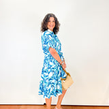 Woman in a blue floral dress holding a beige handbag against a white wall.