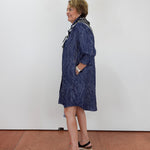 A woman wearing a navy blue patterned long-sleeve shirt dress with a white and black patterned collar, standing in profile against a white wall on a terracotta tiled floor.