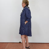A woman wearing a navy blue patterned long-sleeve shirt dress with a white and black patterned collar, standing in profile against a white wall on a terracotta tiled floor.