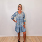 Woman wearing a blue floral dress standing against a white wall.