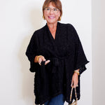 Woman wearing a black poncho and holding a leopard print handbag against a white wall.