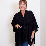 Woman wearing a black poncho and holding a leopard print handbag against a white wall.