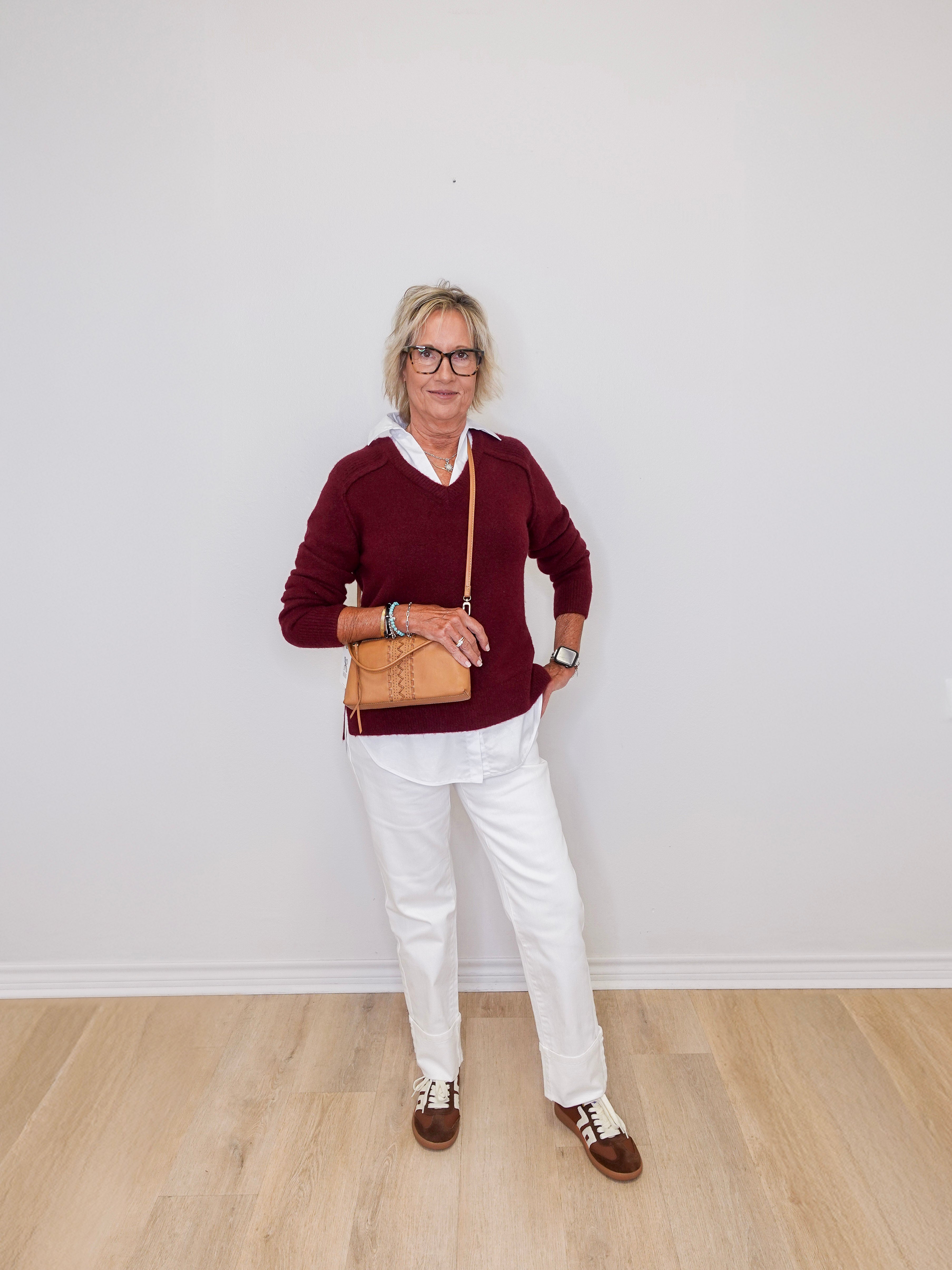Person wearing a burgundy sweater, white pants, and brown shoes against a plain white wall.