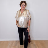 Woman wearing a gold metallic blouse and black pants against a white wall.