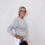 Woman wearing a gray sweater and white pants, holding a black handbag against a white background