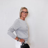 Woman wearing a gray sweater and white pants, holding a black handbag against a white background