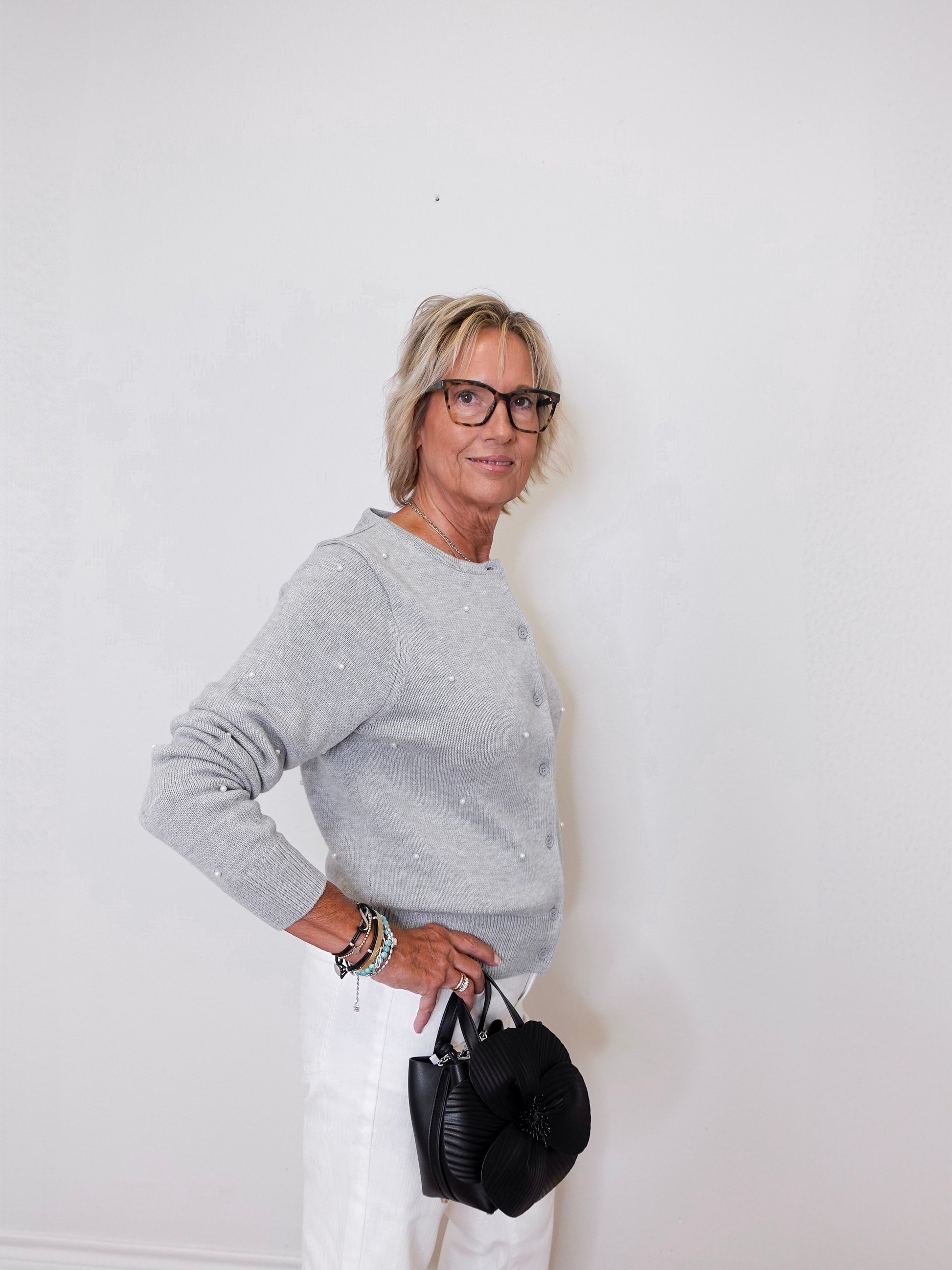 Woman wearing a gray sweater and white pants, holding a black handbag against a white background