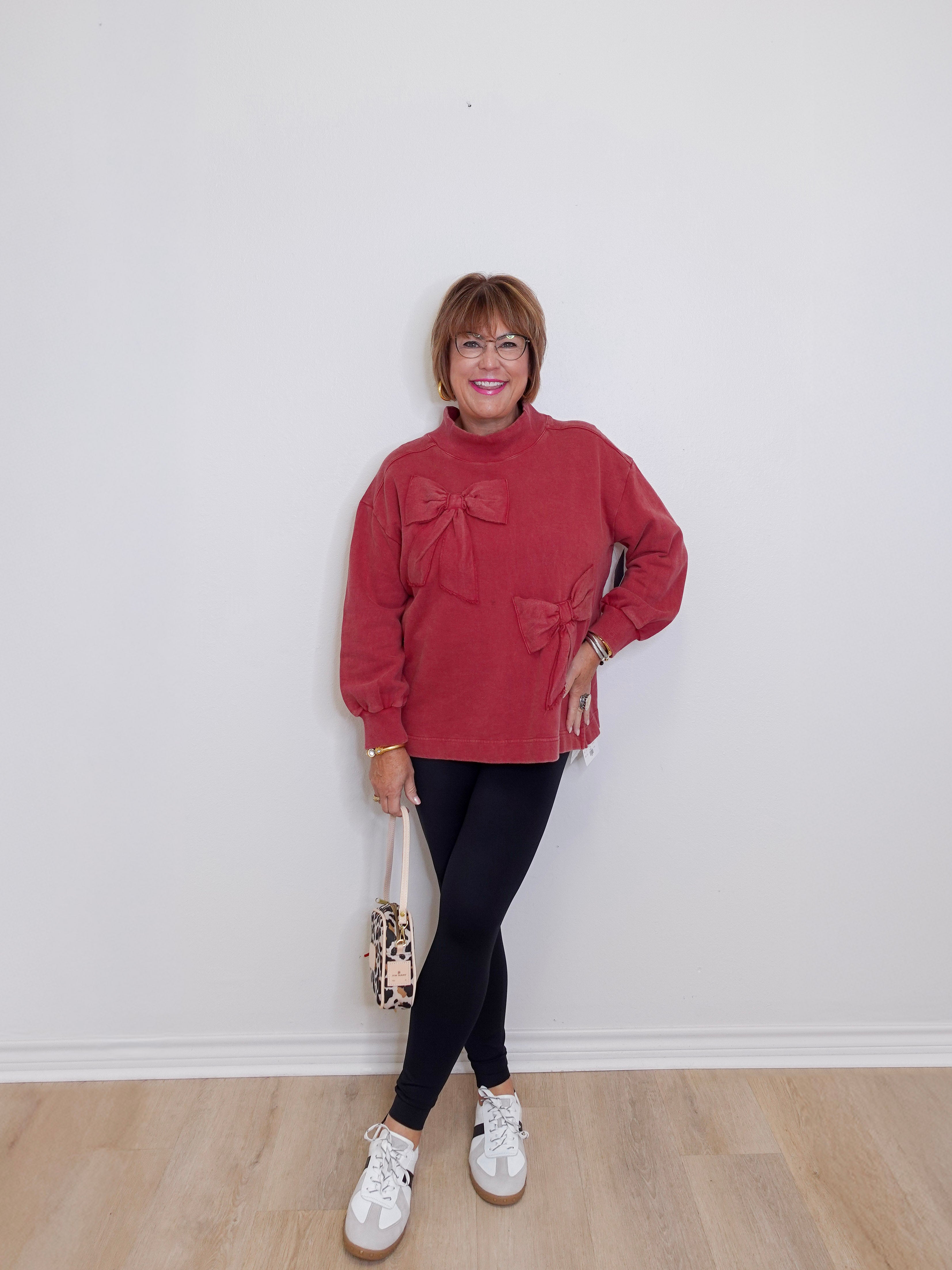 Woman wearing a red sweater with a bow, black pants, and white sneakers against a white wall.