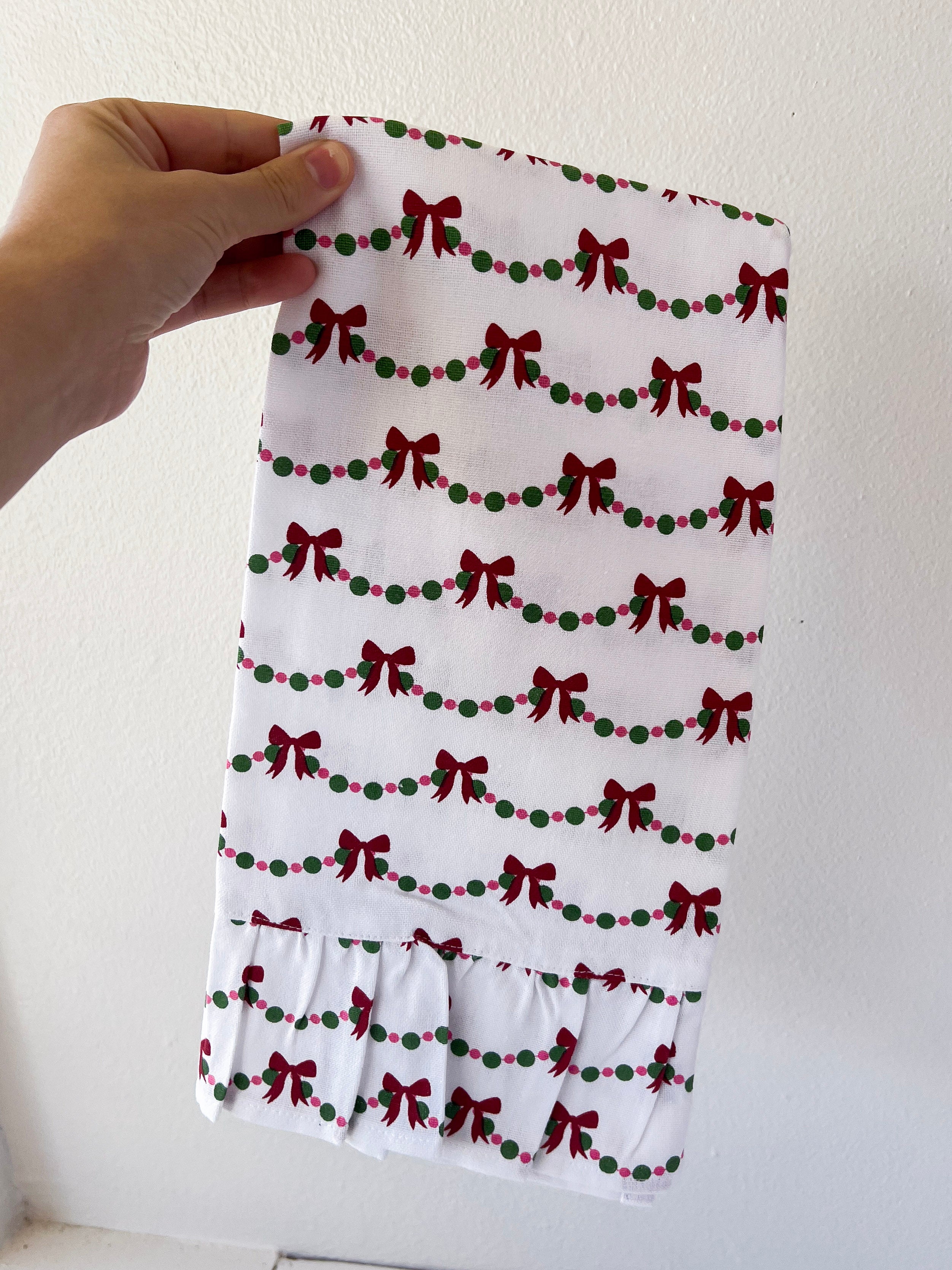 Hand holding a white fabric with red bow and green berry pattern against a plain background