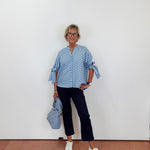 Woman wearing a blue polka dot blouse and black pants, holding a blue handbag against a white wall.
