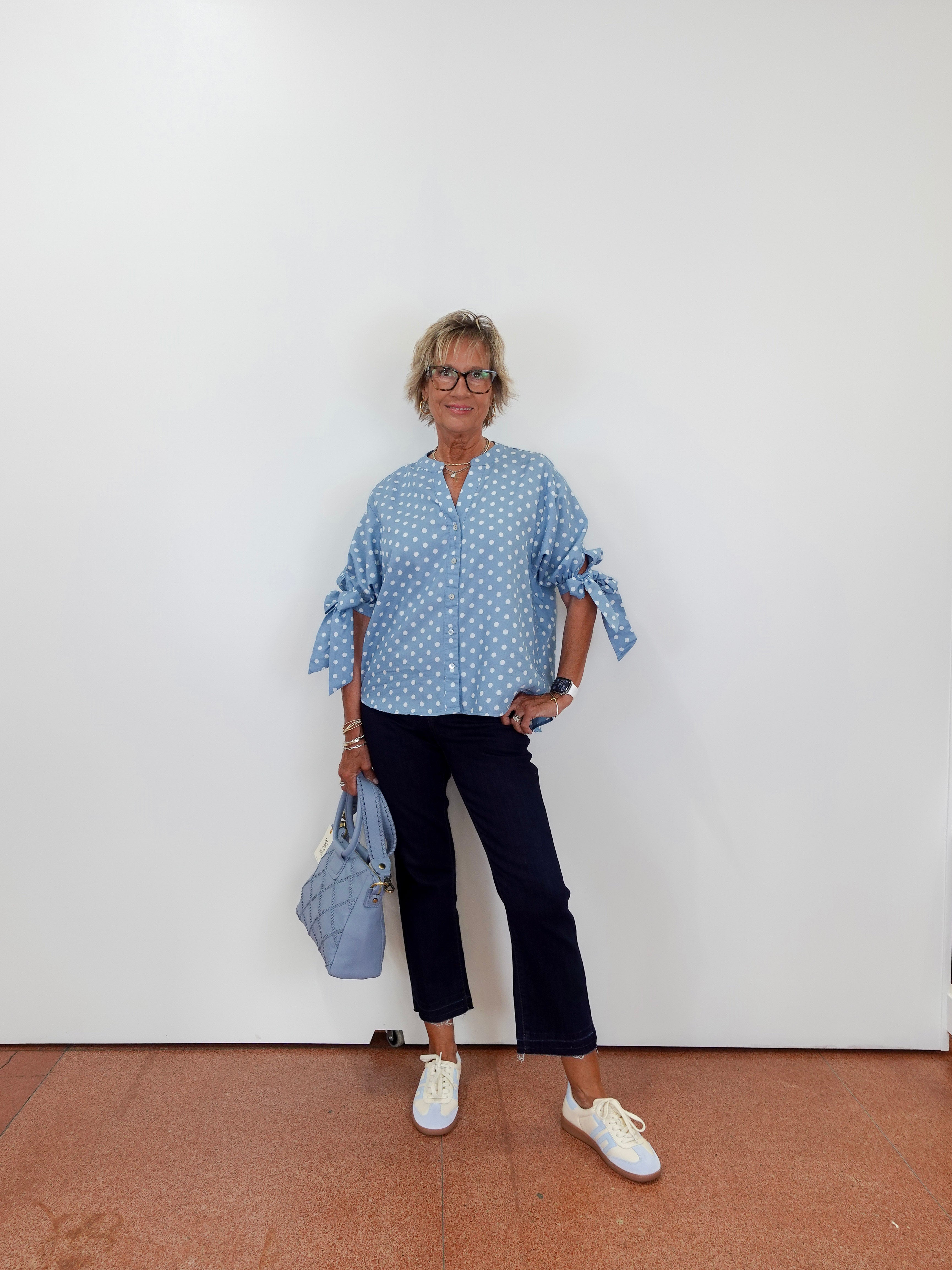 Woman wearing a blue polka dot blouse and black pants, holding a blue handbag against a white wall.