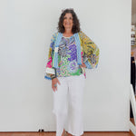 Woman wearing a colorful floral jacket and white pants standing against a white wall.
